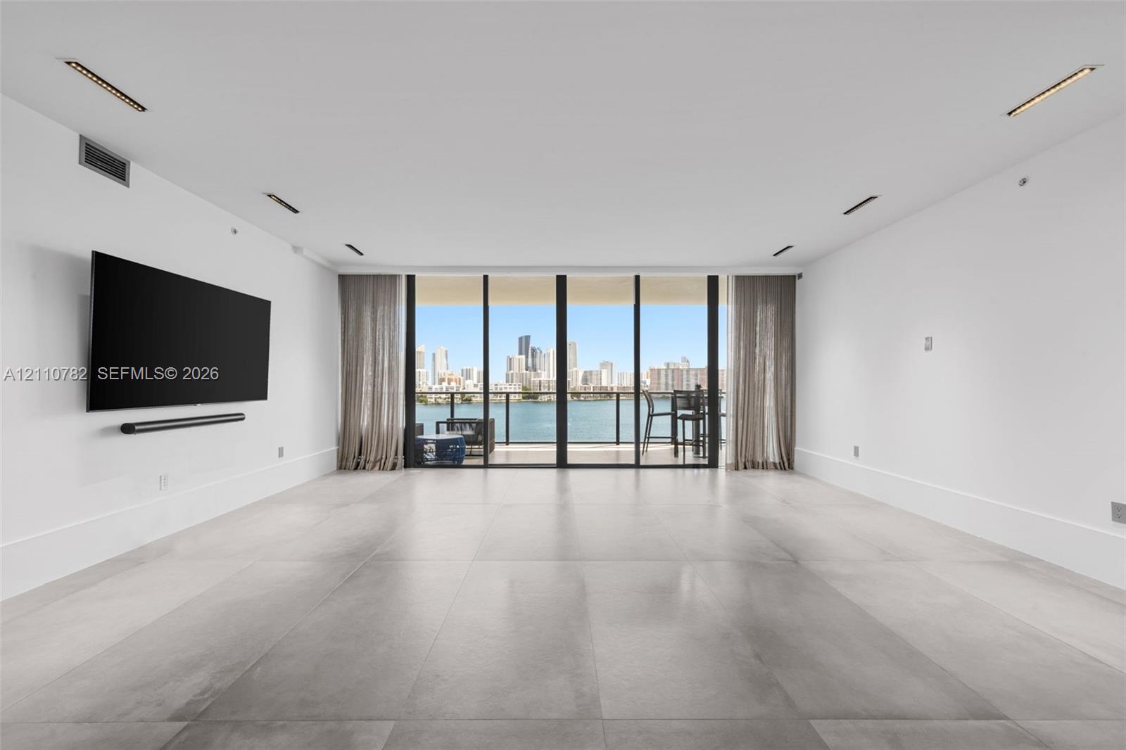 Large-format tile flooring, recessed linear lighting, and expansive floor-to-ceiling glass paneling leading to a balcony with water and skyline scenery