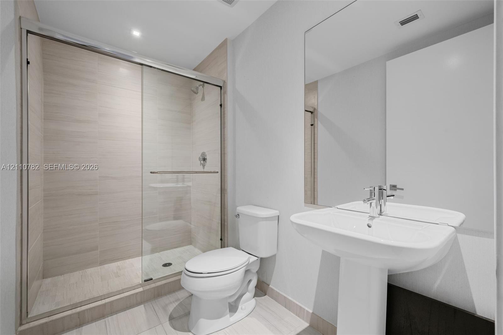 Walk-in shower with sliding glass doors, neutral tile surround, pedestal sink, chrome fixtures, and wall-mounted mirror
