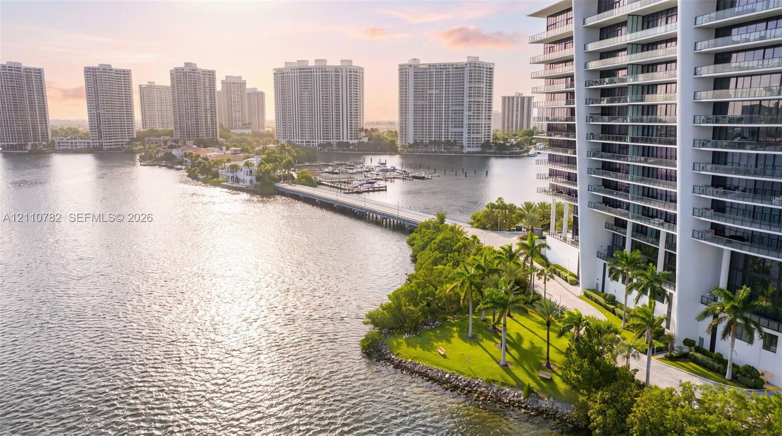 High-rise structure featuring stacked glass-railing balconies, situated on a waterfront lot with manicured lawn, mature palm trees, and direct bulkhead water frontage