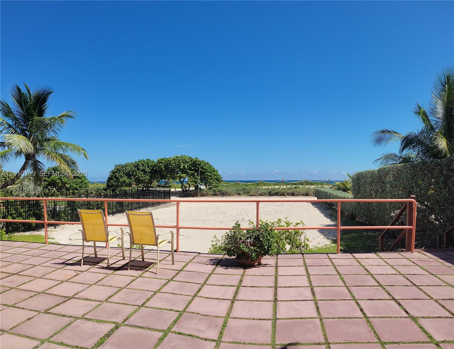 Paved patio featuring a sandy area, metal perimeter fencing, mature palm trees, and coastal scenery