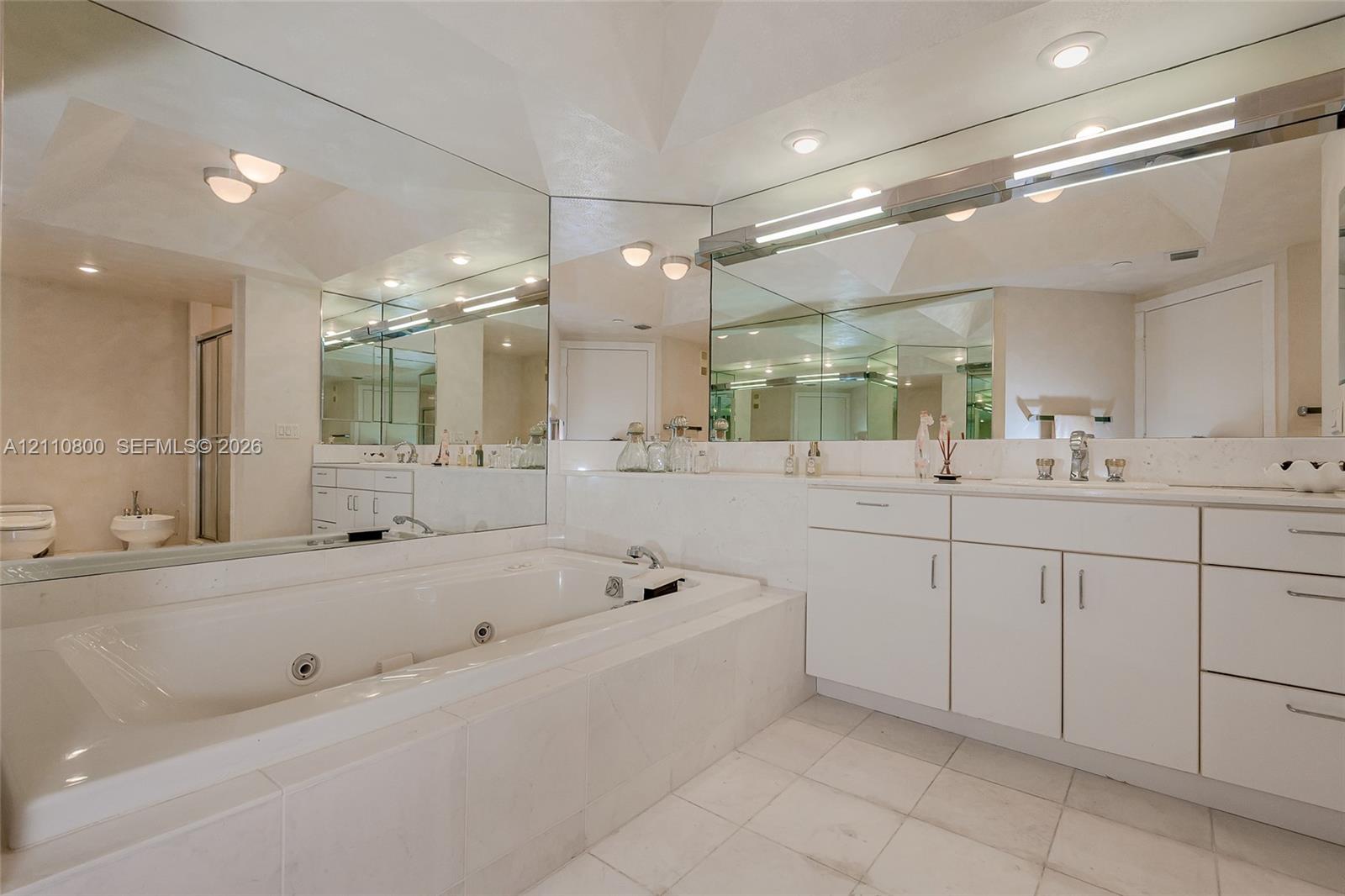 Bathroom featuring a large jetted soaking tub with a tile surround, white cabinetry, expansive wall-to-wall mirrors, recessed lighting, and tile flooring
