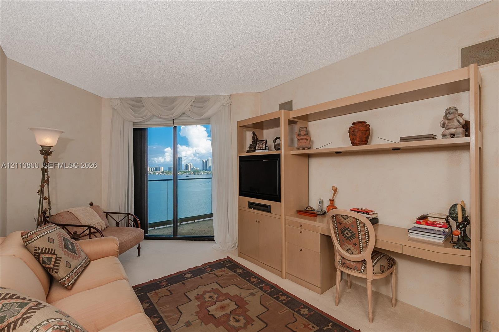 Built-in light-tone wood cabinetry featuring open shelving and a dedicated workstation, floor-to-ceiling glass sliding door, and private balcony with water backdrop