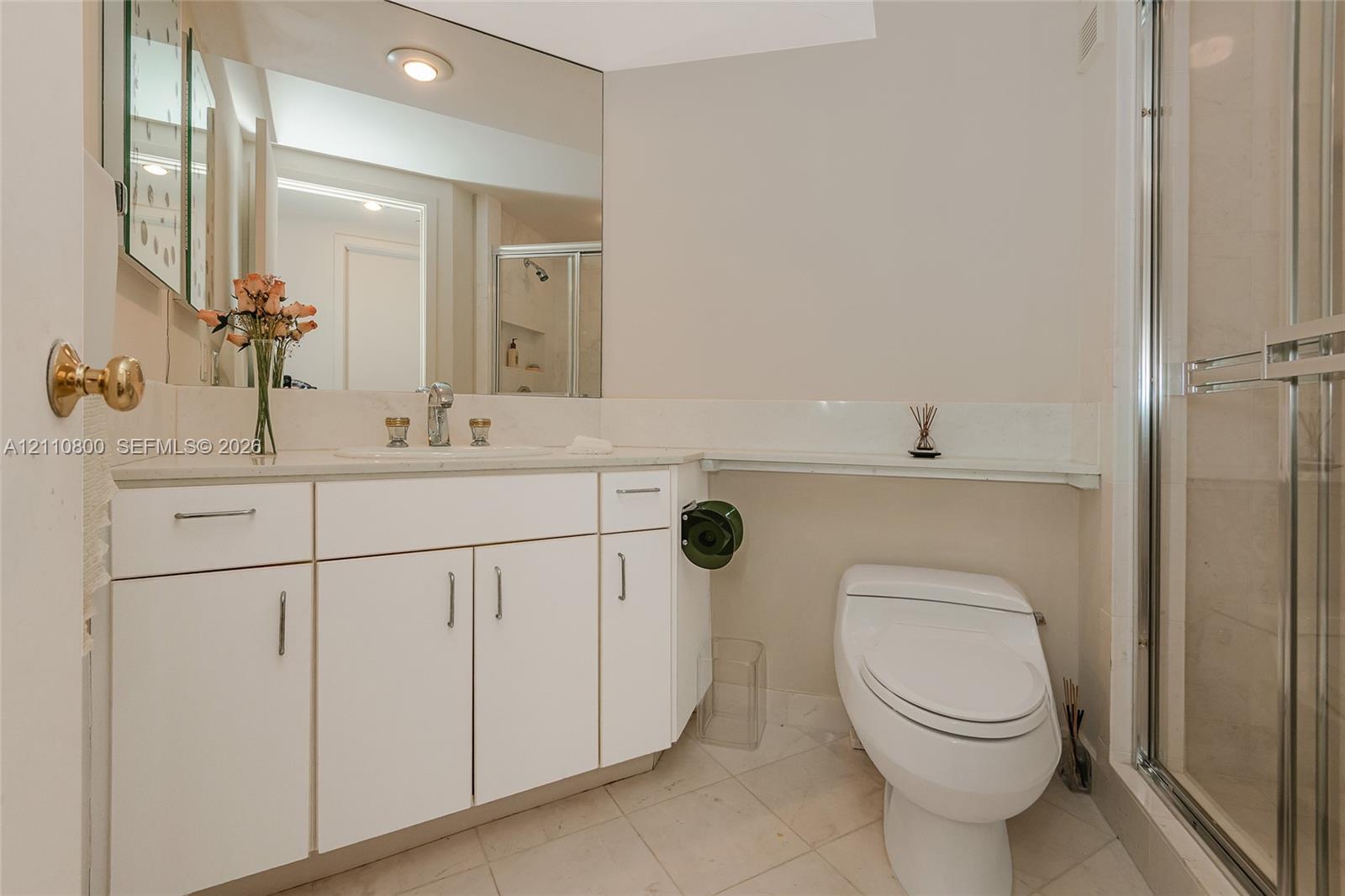 Bathroom featuring white cabinetry, an integrated basin with a chrome faucet, wall-spanning mirror, elongated toilet, and a glass-enclosed shower