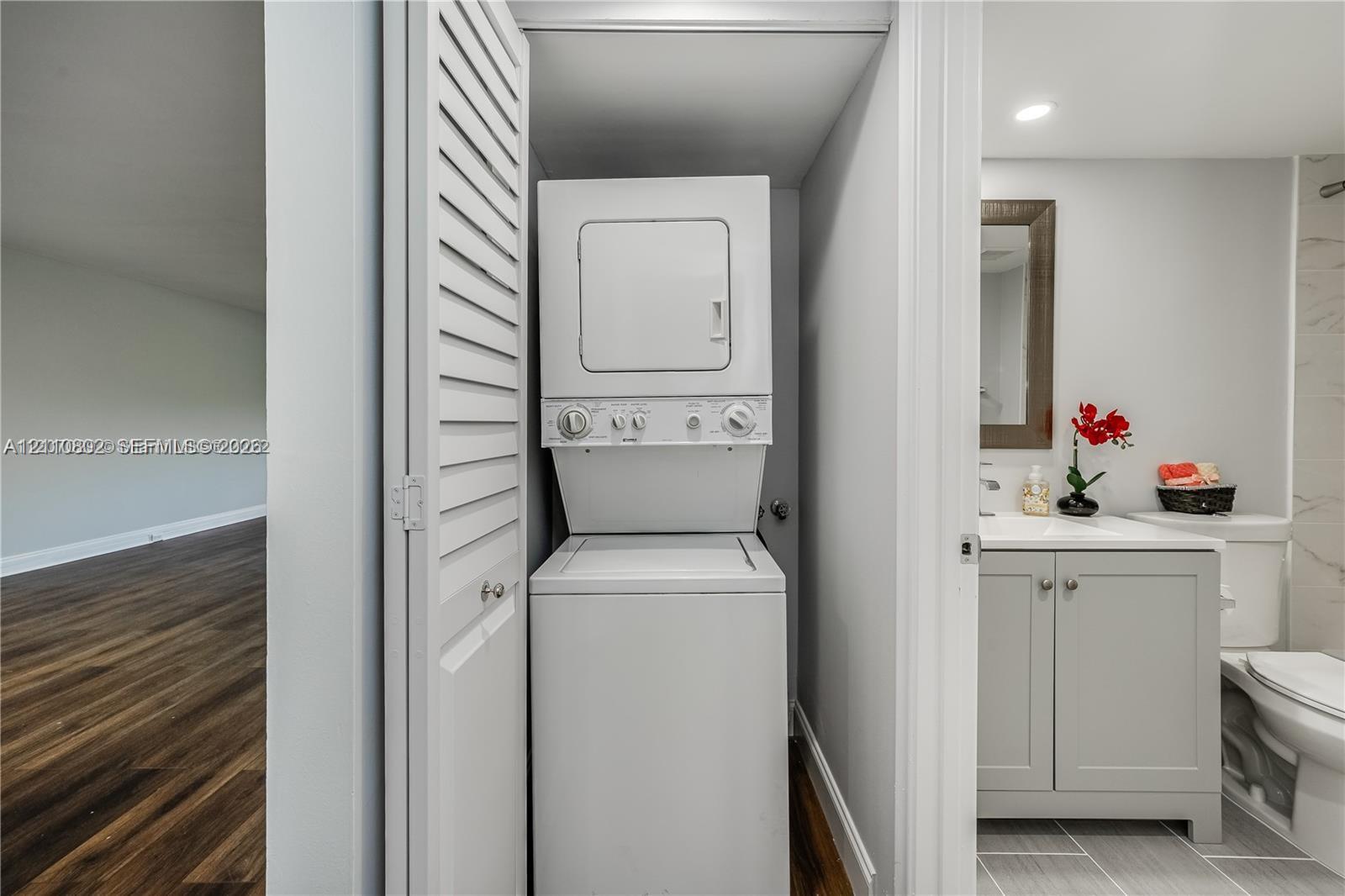 Laundry closet featuring a white stacked washer and dryer unit behind louvered bi-fold doors, grey vanity cabinet with a white countertop, and tiled flooring