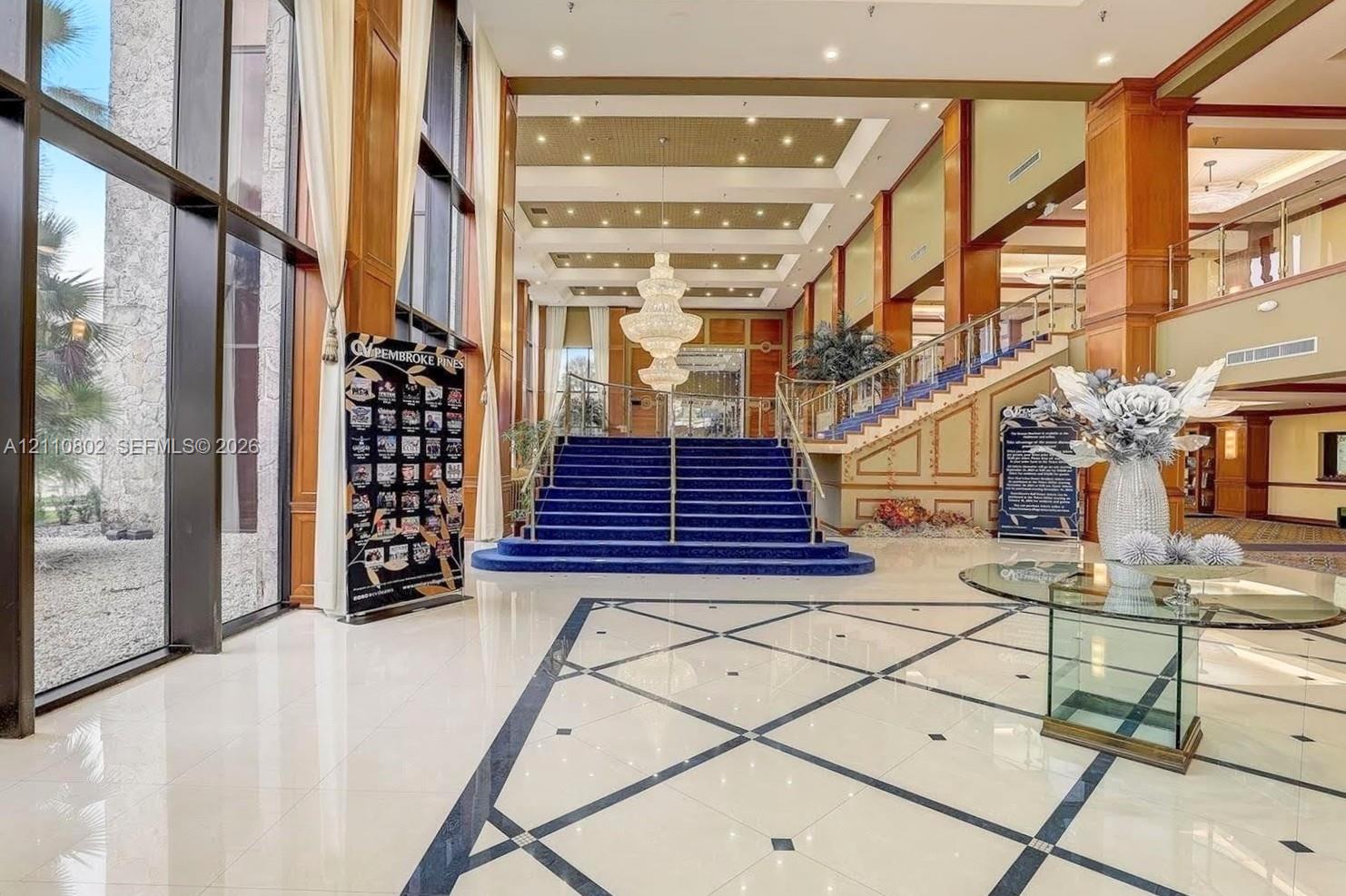 Grand foyer featuring expansive floor-to-ceiling windows, polished stone tile flooring with decorative inlay, a central multi-tier crystal chandelier, recessed coffered ceilings, and a staircase with an ornamental metal railing