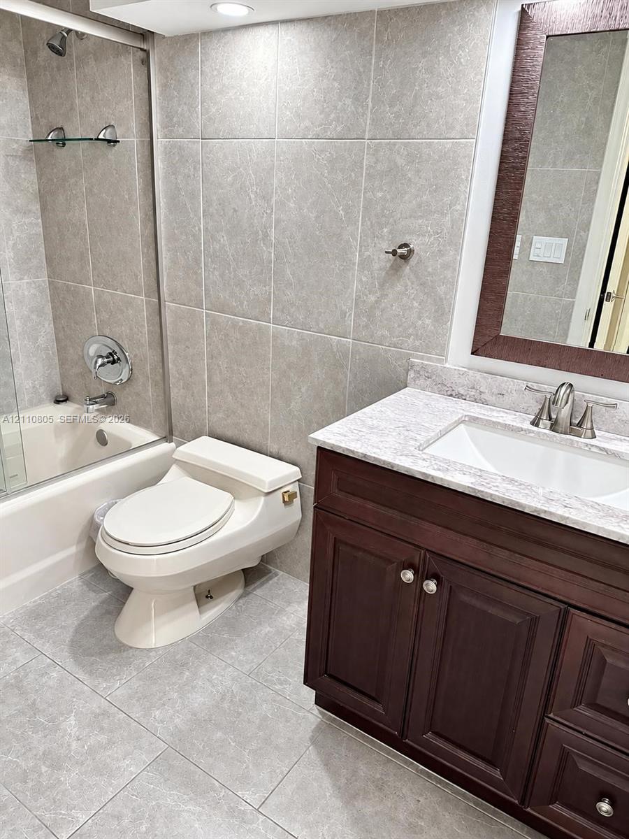 Bathroom featuring a dark wood-finish vanity with a stone-composite countertop, full-height tile wall surrounds, glass shower enclosure, and oversized ceramic floor tiling