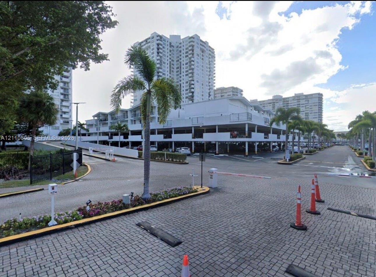 Covered parking structure featuring multiple levels, paved brick driveway, and manicured landscaping with palm trees