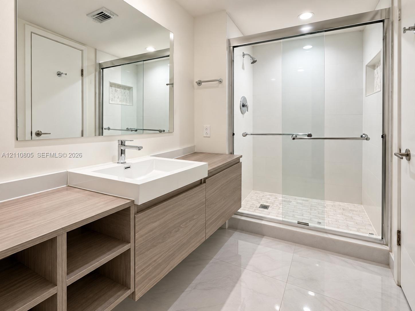 Floating vanity featuring a wood-finish cabinet with integrated shelving, rectangular vessel sink, polished chrome faucet, and glass-enclosed shower with a tiled floor and built-in niche