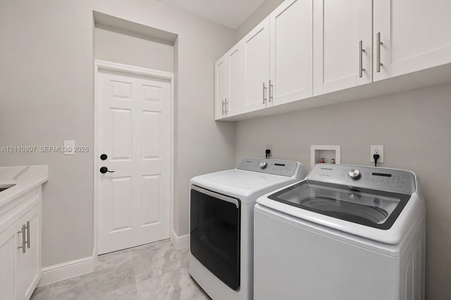 Laundry room featuring white Shaker-style upper cabinetry, stone-finish tile flooring, solid-surface countertop with integrated sink, and matte black door hardware