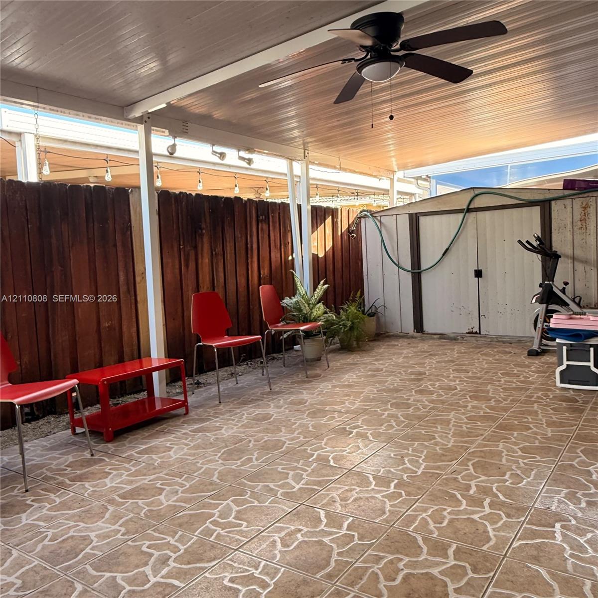 Covered patio featuring patterned tile flooring, wood-panel fencing, a ceiling fan, and a detached metal storage structure
