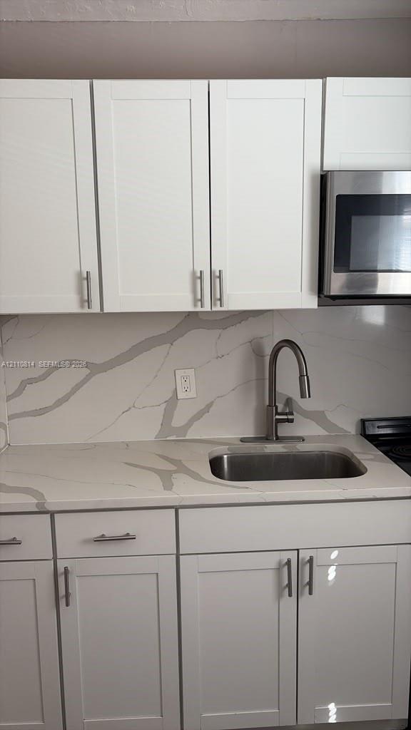 Shaker-style white cabinetry, stone-finish countertops, coordinating full-height backsplash, stainless steel undermount sink with arched faucet, and integrated stainless steel microwave