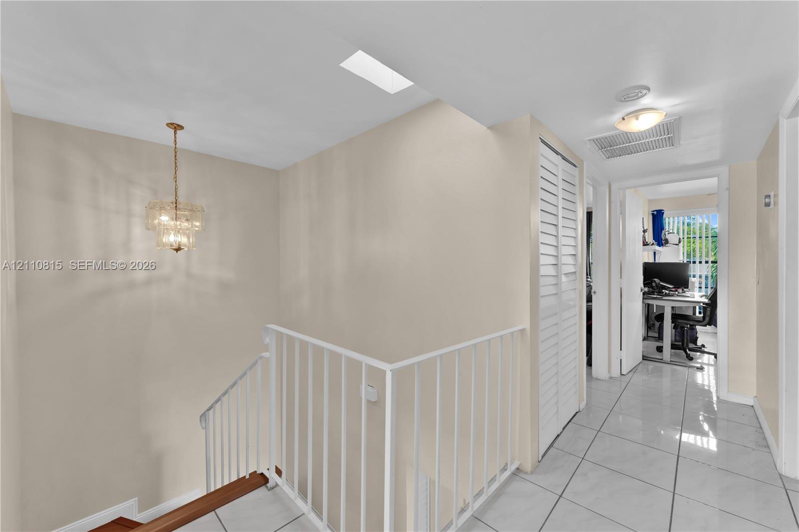 Interior hall featuring white tile flooring, ceiling-mounted crystal chandelier, skylight, and white louvered closet doors with a metal railing