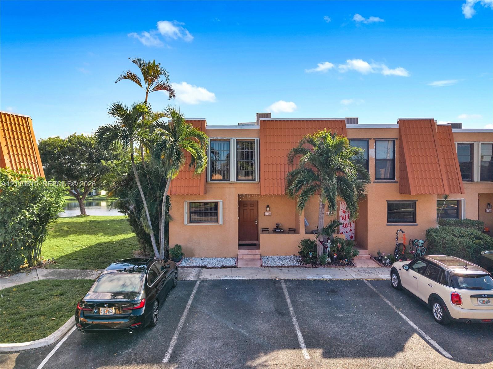 Two-story exterior featuring stucco siding, prominent mansard-style roof elements, dual assigned parking spaces, and mature palm trees