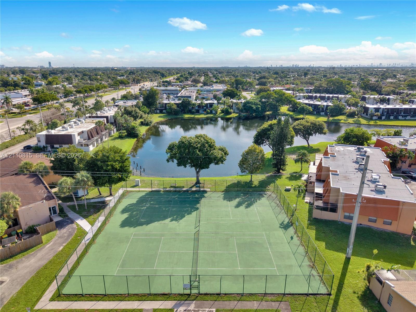 Aerial perspective of a fenced tennis court situated by a waterfront pond featuring mature landscaping and surrounding multi-unit residential structures