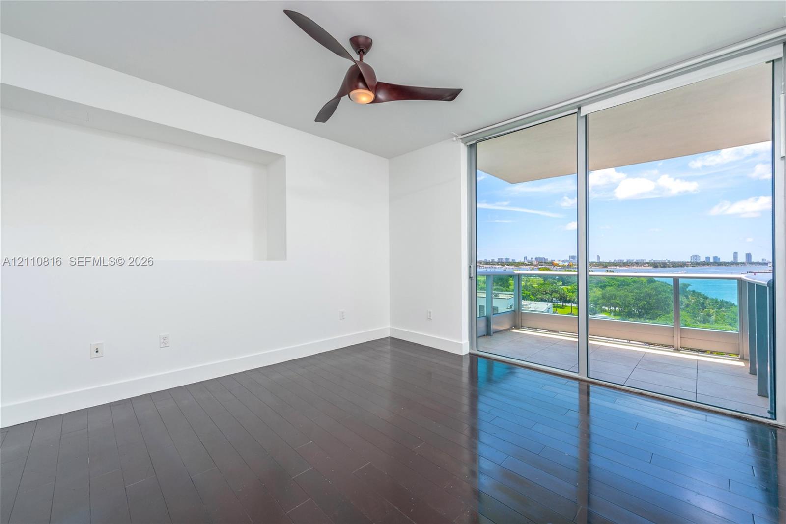 Dark wood-finish flooring, recessed wall niche, modern blade-style ceiling fan, and floor-to-ceiling sliding glass doors opening to a private balcony with glass railing
