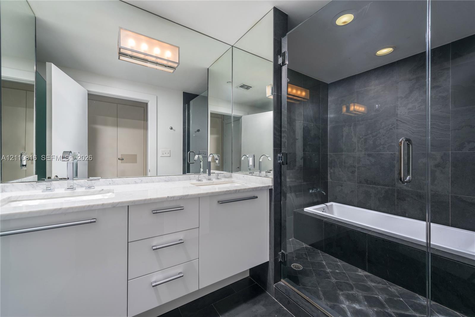 Bathroom featuring white cabinetry, stone-finish vanity countertop, dual undermount sinks with polished chrome fixtures, full-height vanity mirror, and a glass-enclosed bathtub with dark tile surround