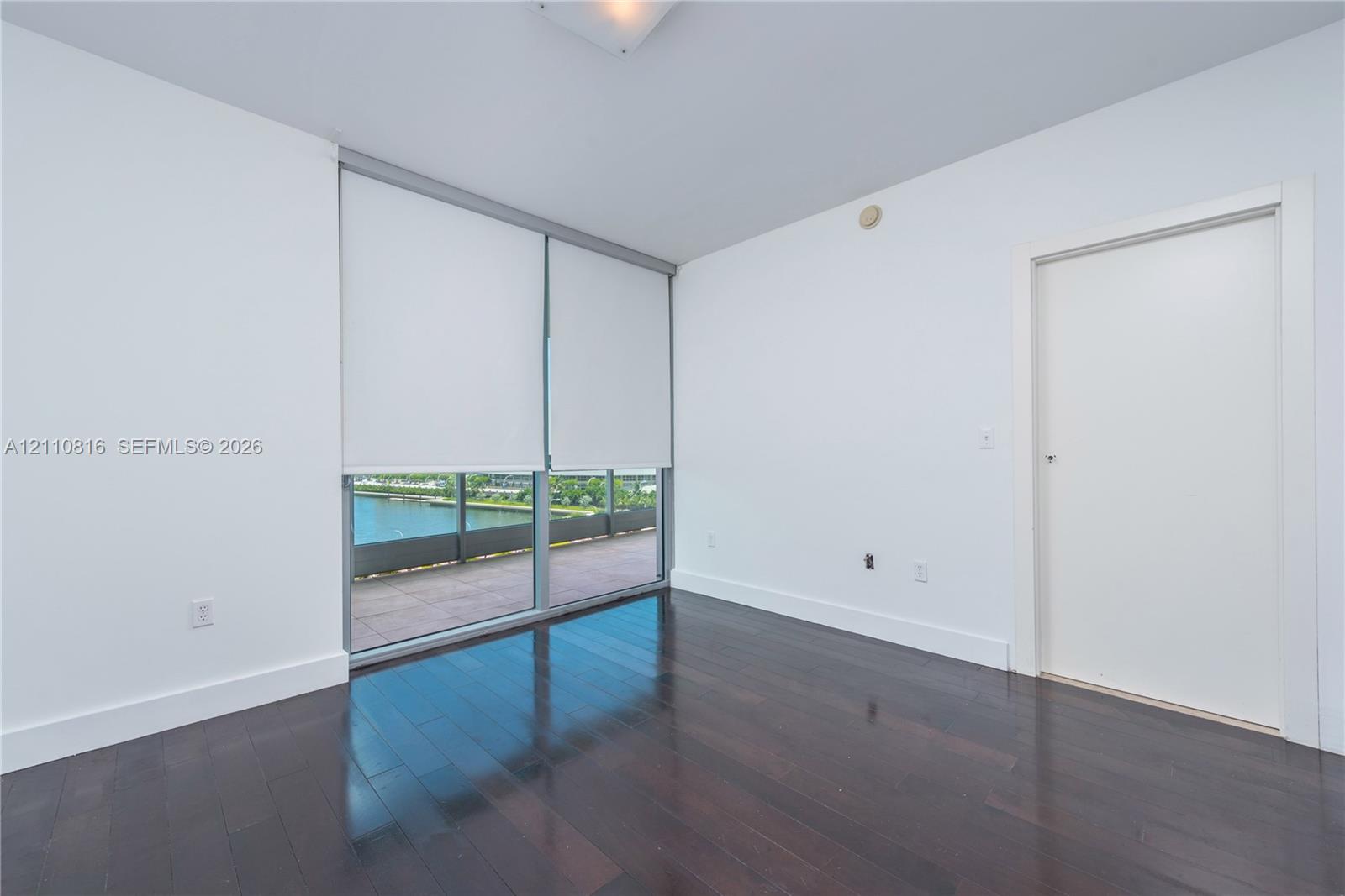 Room featuring dark wood-finish flooring, floor-to-ceiling windows, and access to a balcony with water scenery