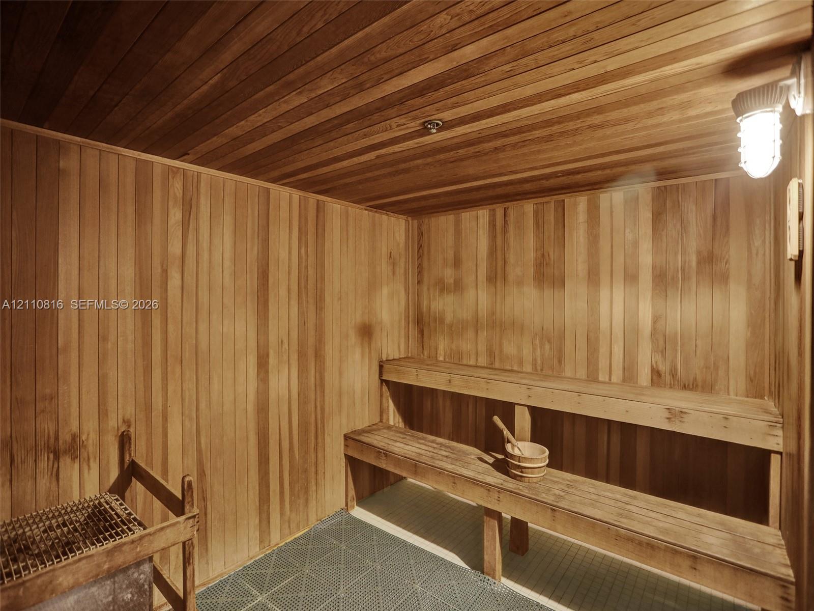 Sauna interior featuring natural wood-paneled walls and ceiling, multi-level tiered wooden benches, an integrated stone heater, and industrial-style wall-mounted lighting