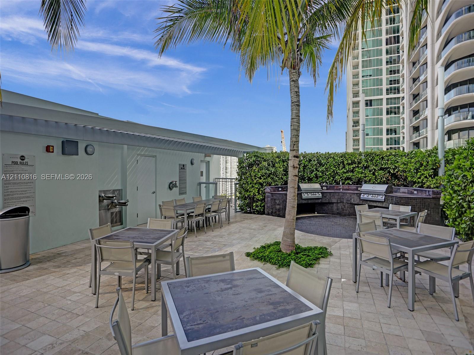 Outdoor terrace featuring stone-tile flooring, a built-in stone-finished kitchen station with integrated stainless steel grills, a manicured hedge perimeter, and palm landscaping