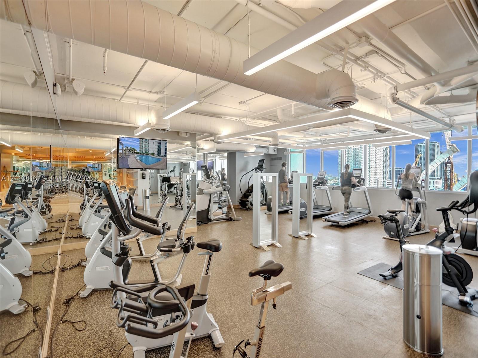 Fitness center featuring industrial-style exposed ductwork, wall-to-wall mirrored surfaces, expansive windows, linear overhead lighting, and stone-finish flooring