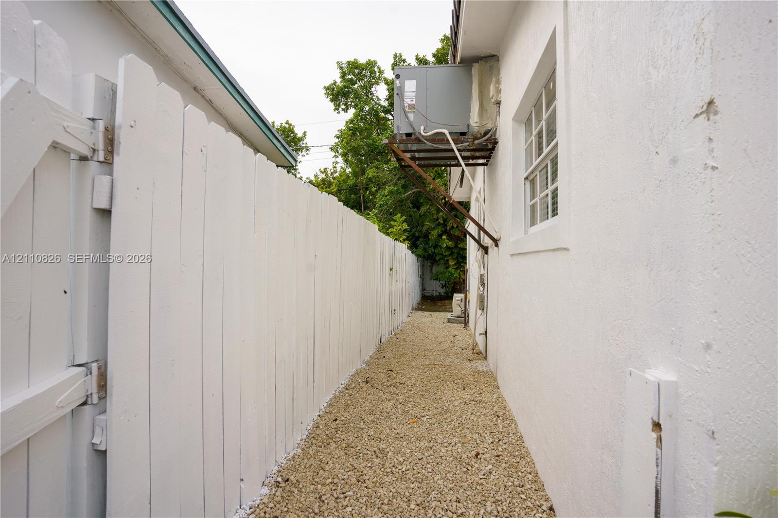 Side exterior area featuring a white picket fence, gravel ground cover, and a wall-mounted HVAC unit