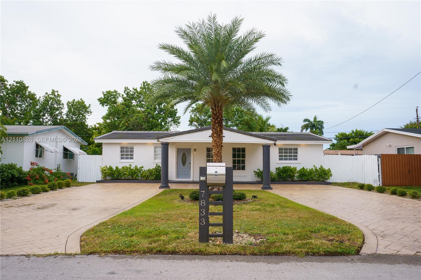 Single-story residence featuring a covered entryway with structural columns, dual paved driveways, a central palm tree, and white privacy fencing
