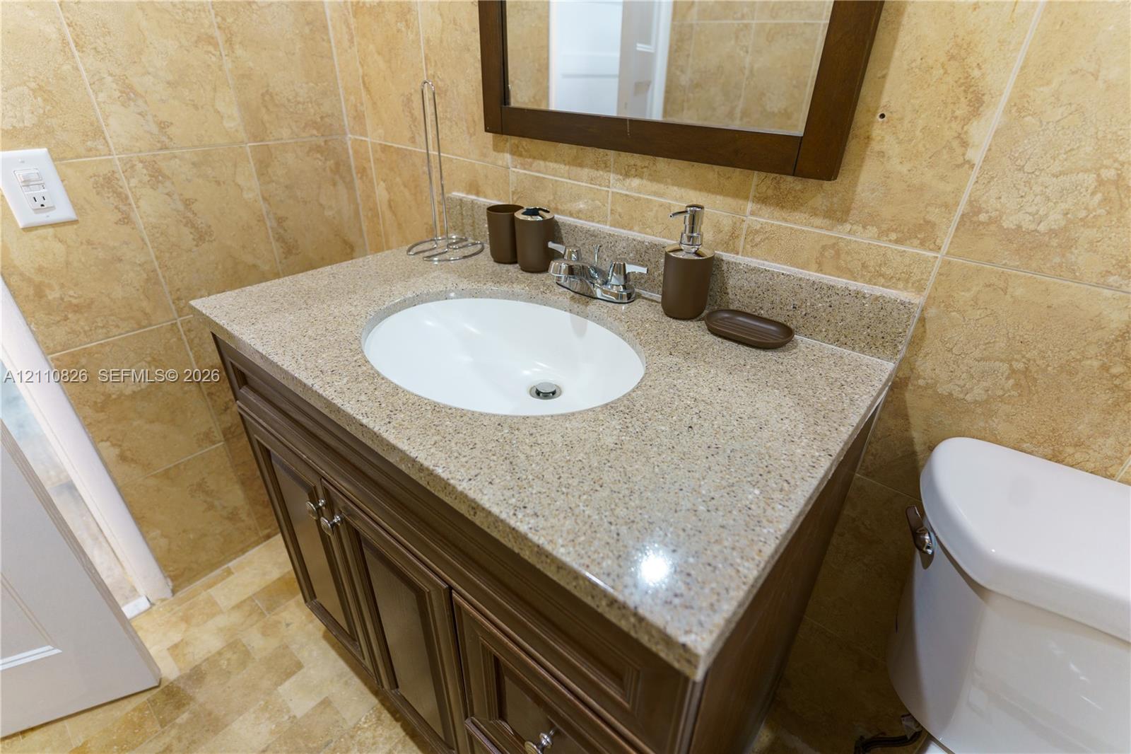 Vanity featuring a stone-composite countertop with an integrated oval sink, dark wood cabinetry, and a framed mirror surrounded by neutral-toned tiled walls