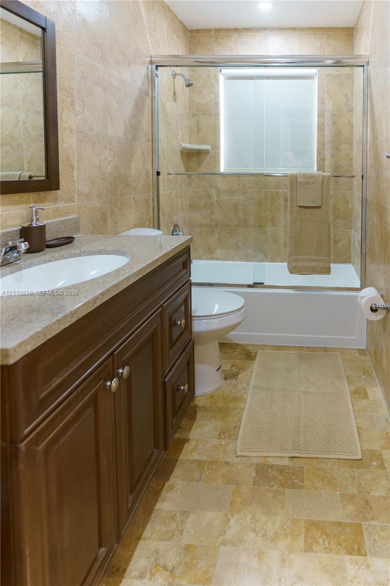 Bathroom featuring a wood-finish vanity with a stone-composite countertop, full-height wall tiling, and a tub-shower combination enclosed by a glass sliding door