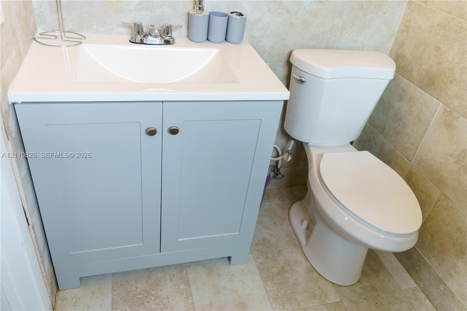 Light gray shaker-style vanity with integrated sink, chrome dual-handle faucet, and large-format neutral tile flooring and wall surfacing