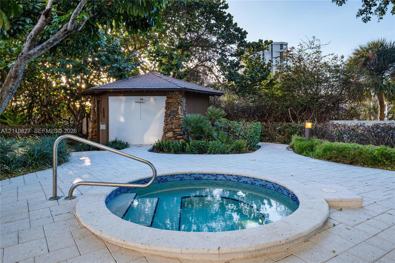 In-ground spa with tile detailing, stone-accented cabana, integrated metallic handrail, and light-toned paver hardscaping