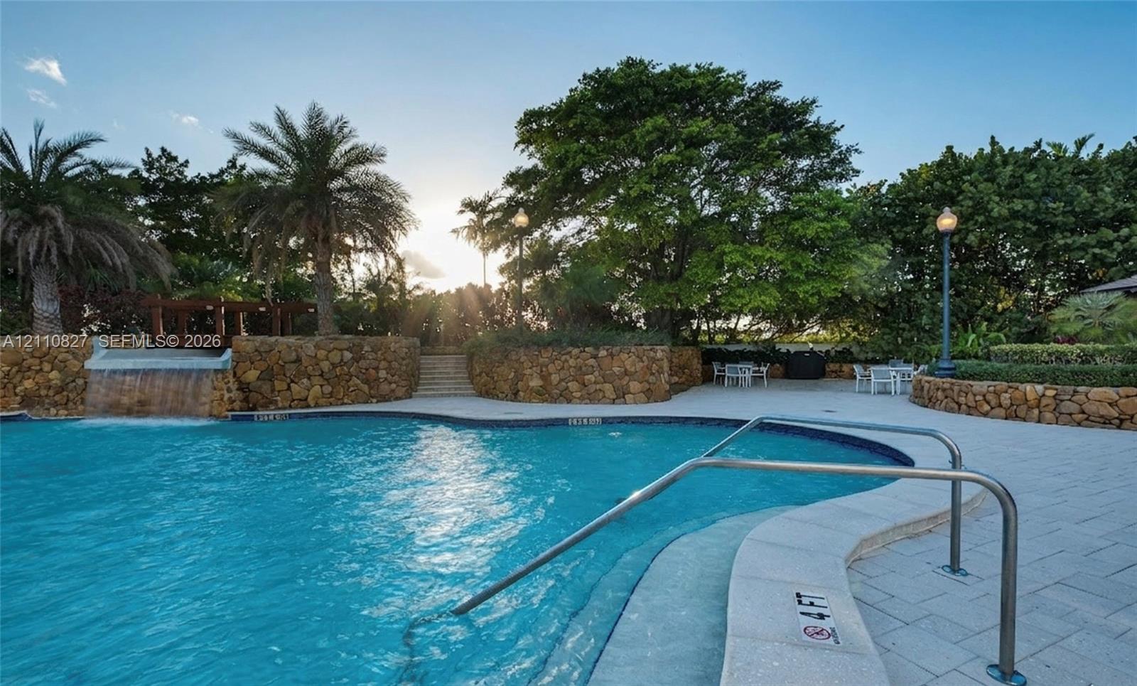 Swimming pool featuring a rock-faced water feature, paver-stone decking, metal handrails, and surrounding mature landscaping