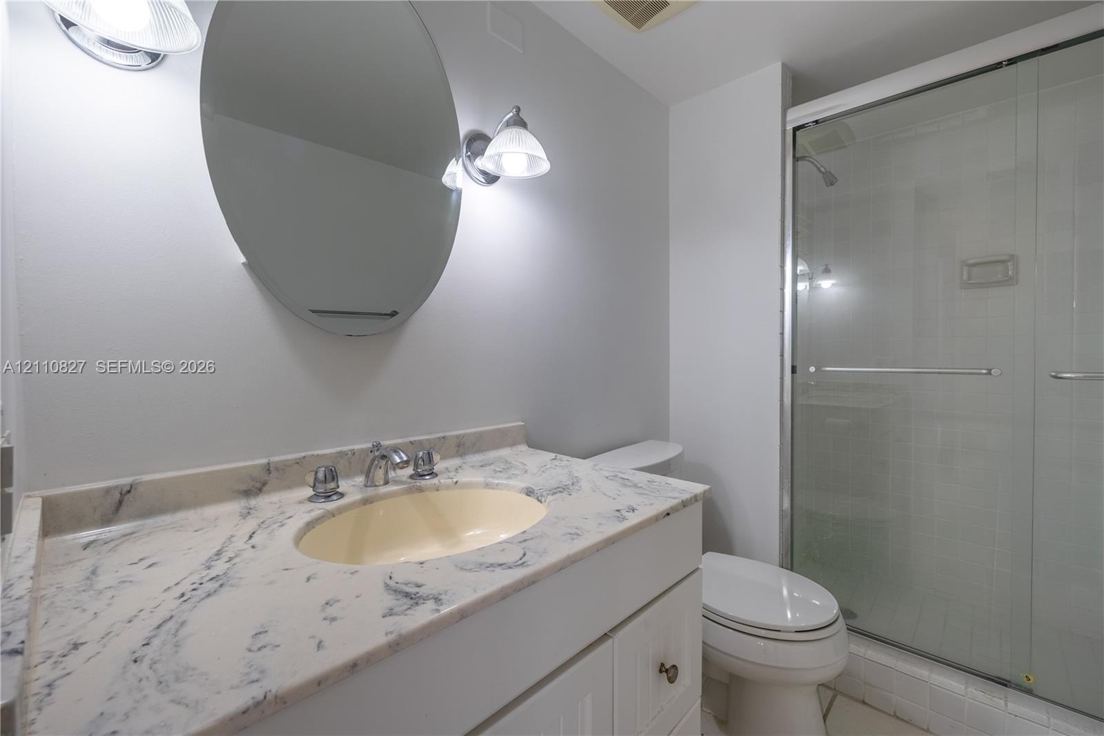 Stone-surfaced vanity with an integrated oval sink, oval wall-mounted mirror, dual-light fixture, and glass-enclosed shower stall