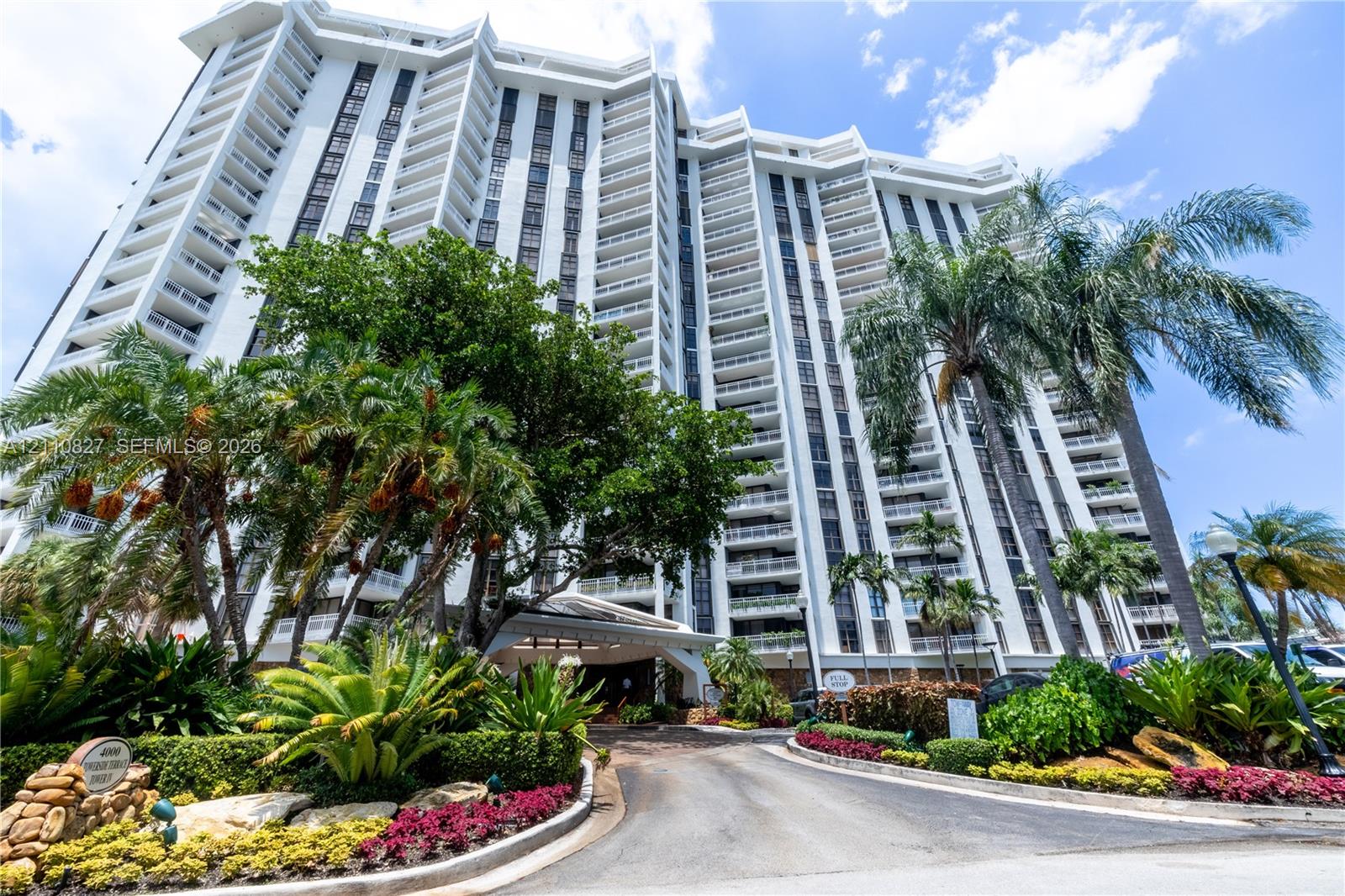 High-rise residential exterior featuring stepped architectural balconies, white concrete facade, covered porte-cochere entry, and extensive tropical landscaping