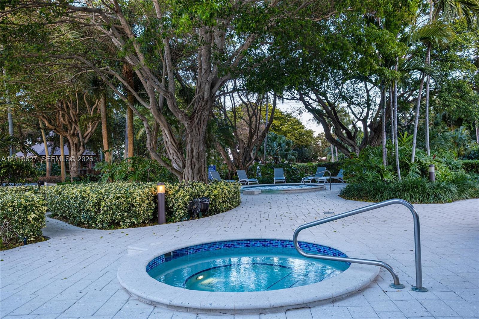 In-ground spa with mosaic tile interior and integrated metal handrail, situated on a stone-paver deck surrounded by mature landscaping