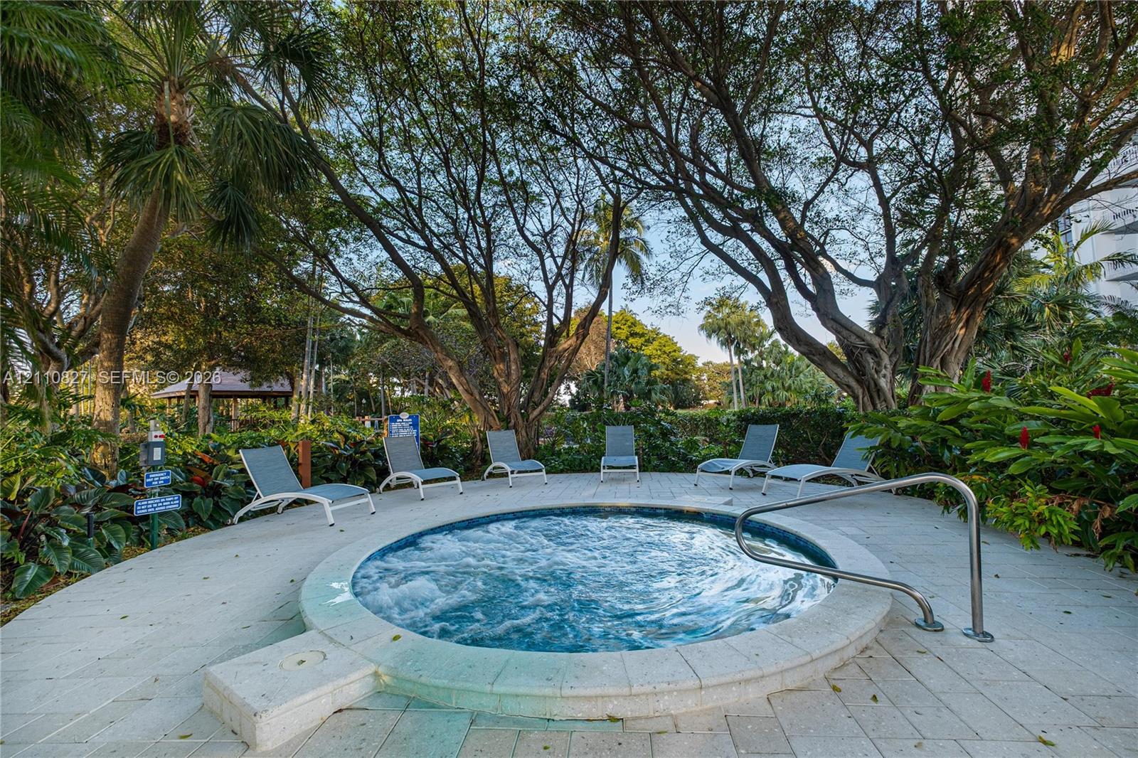 In-ground circular spa featuring a stone paver deck, stainless steel handrail, and mature tropical landscaping