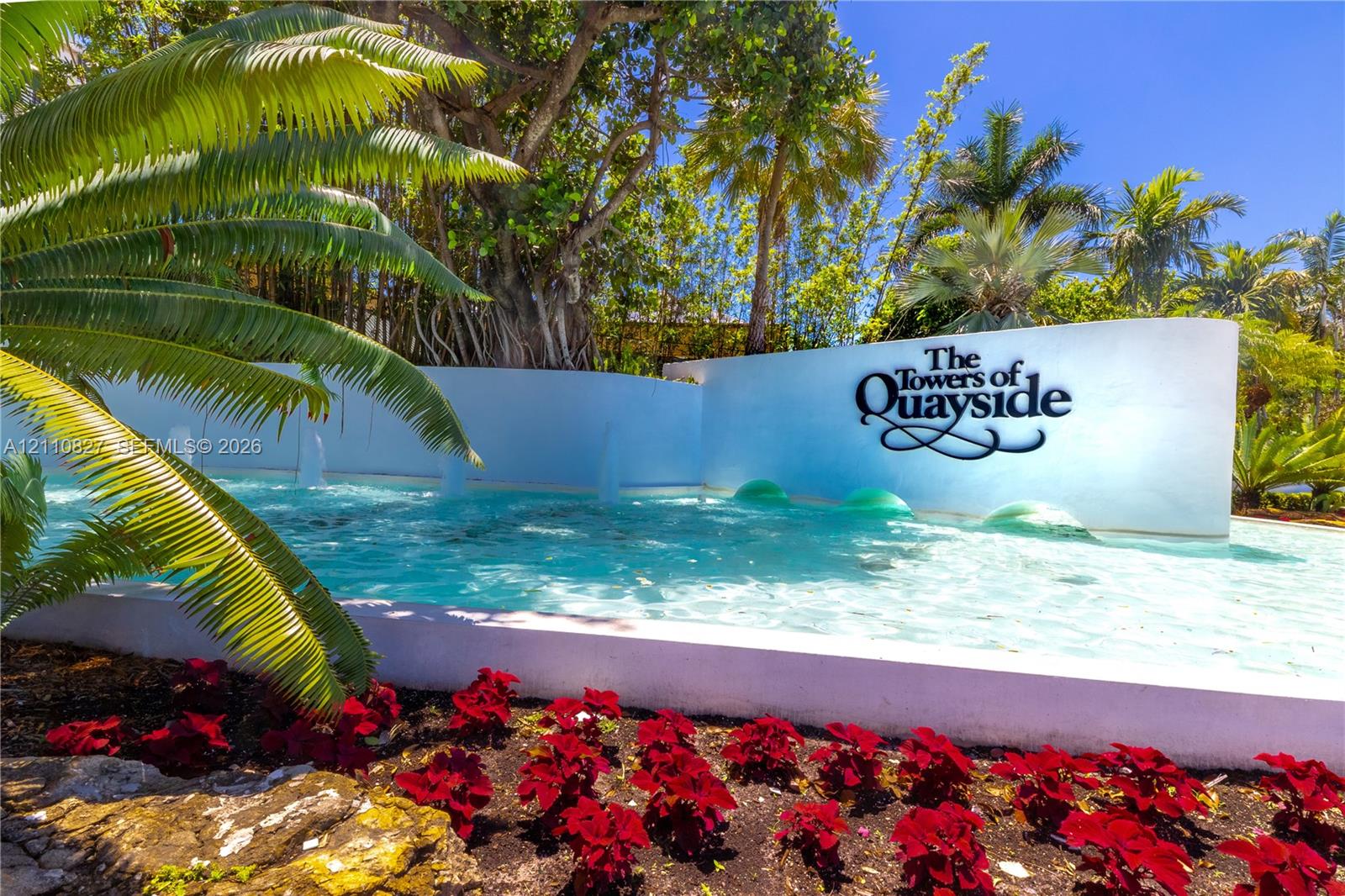 Exterior entrance featuring a water fountain, white masonry retaining wall, mature palm trees, and landscaped floral beds