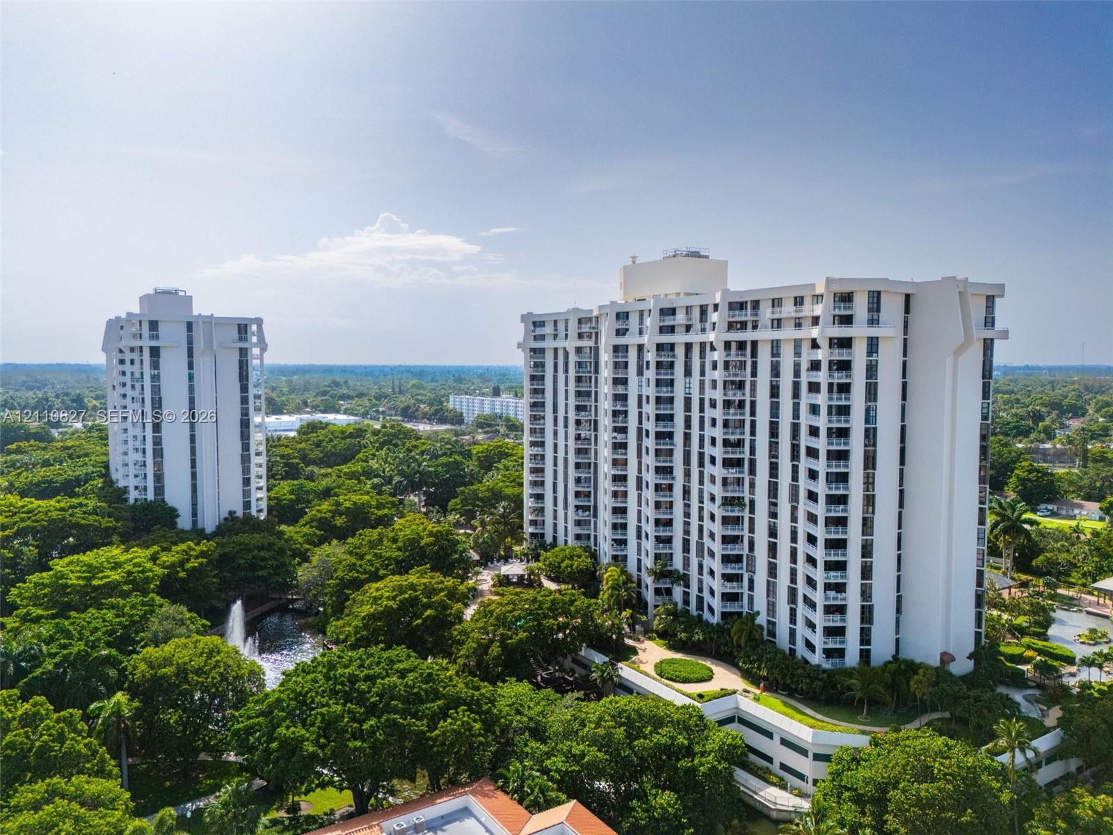 High-rise residential towers surrounded by mature trees, landscaped grounds, pedestrian walkways, and a decorative water feature