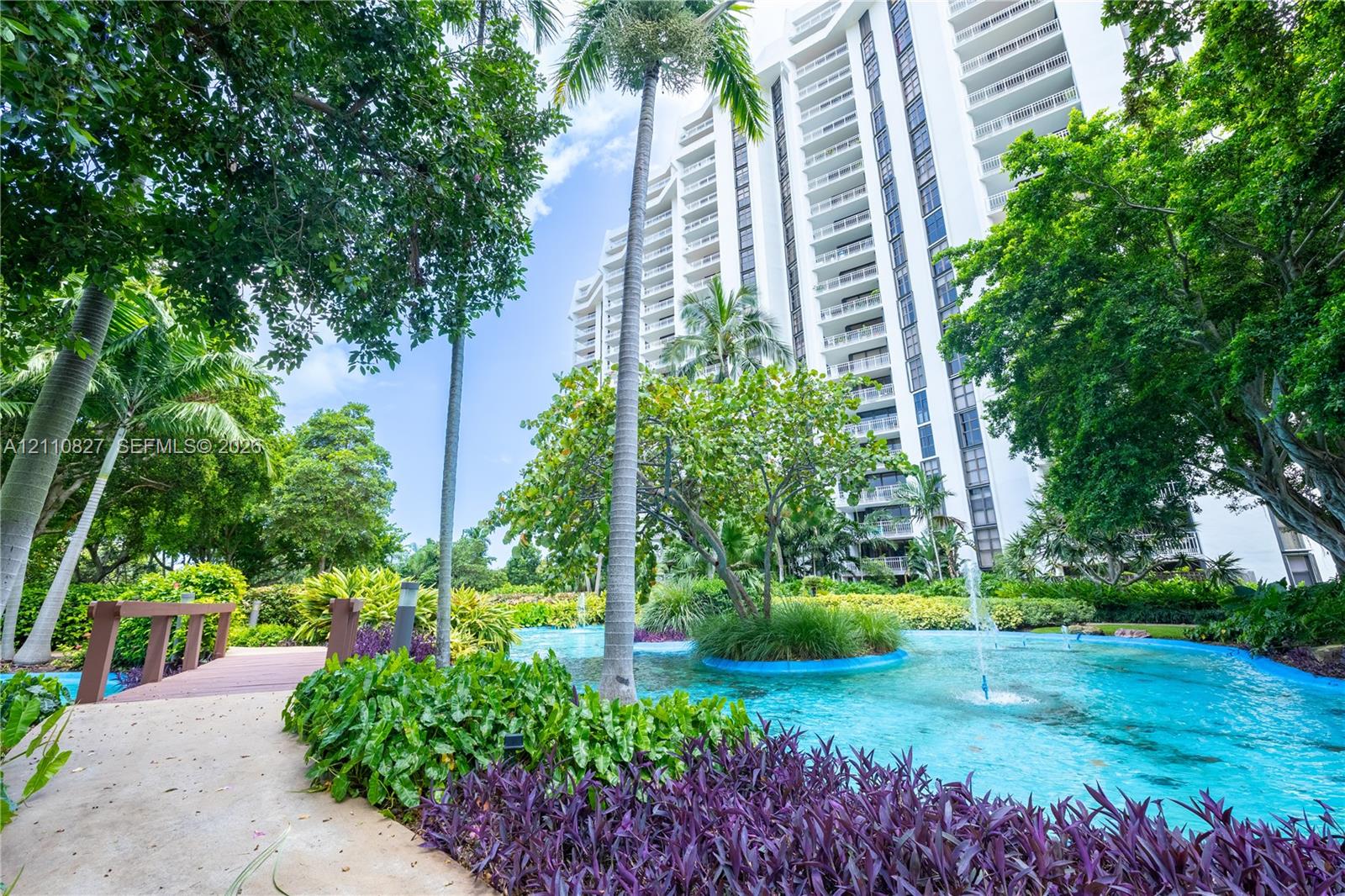 Multi-story high-rise building featuring a landscaped courtyard with a decorative pond, integrated fountains, mature palm trees, and a pedestrian footbridge