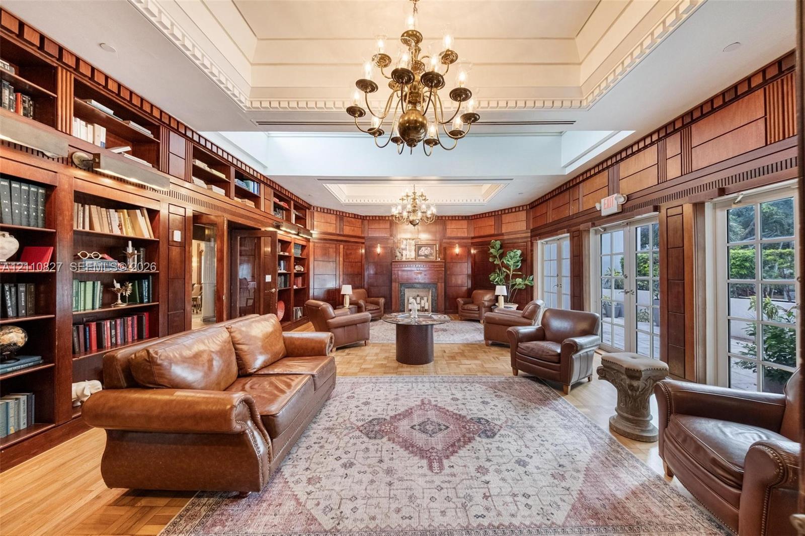 Extensive floor-to-ceiling wood paneling, built-in library shelving, dual tiered tray ceilings with decorative millwork, multiple sets of glass-paneled doors, and a central fireplace with a wood mantel