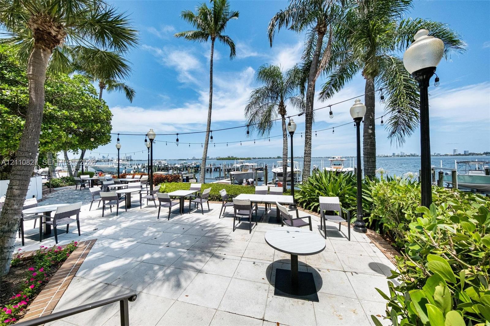 Waterfront patio featuring stone paver flooring, mature palm trees, perimeter garden beds, and post-mounted lighting