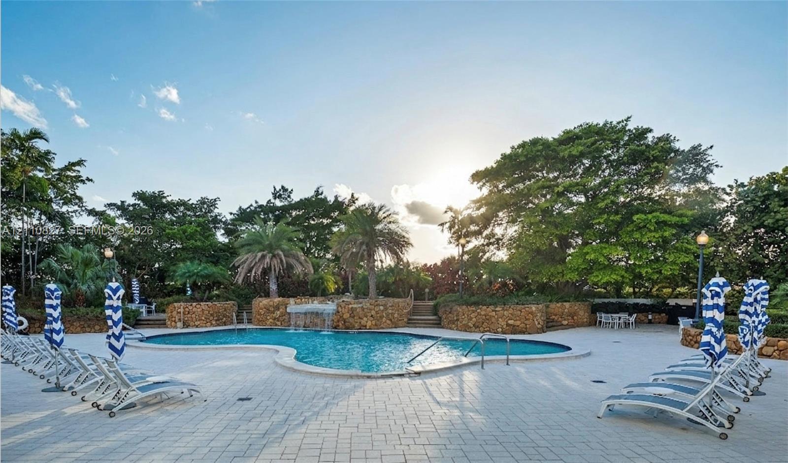 Free-form swimming pool featuring a waterfall feature and stone masonry retaining walls, surrounded by a light-toned paver deck and mature landscaping