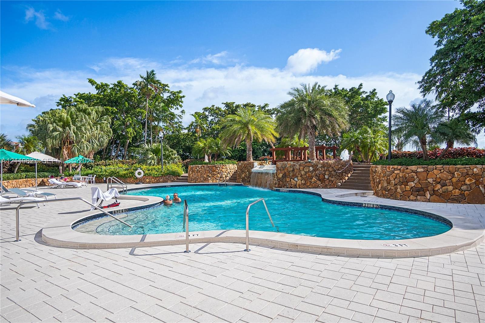 Freeform pool with a water feature, stone retaining walls, paved deck, and mature palm landscaping