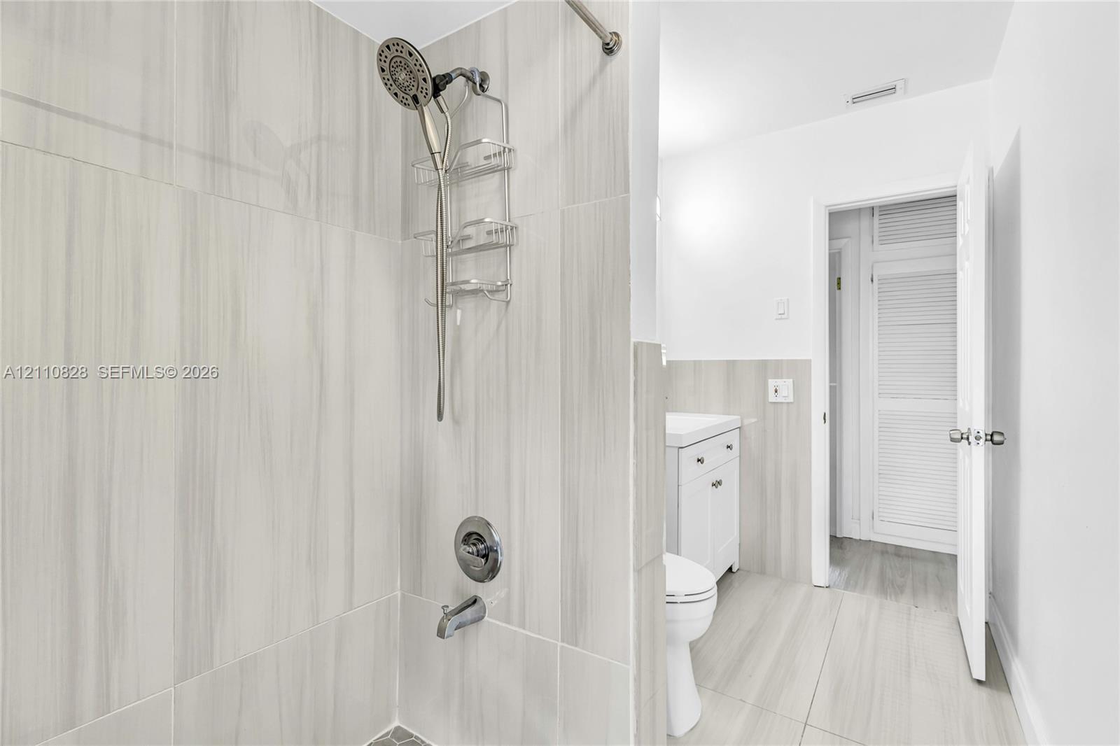 Bathroom featuring neutral large-format tile surround, chrome shower hardware, white vanity cabinetry, and louvered closet doors