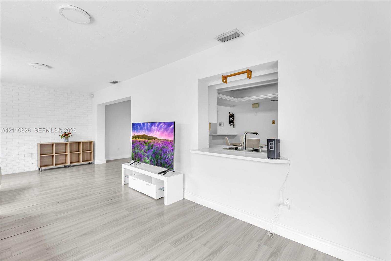 Interior space featuring light-toned wood-finish flooring, white painted brick accent wall, and a kitchen pass-through window with a countertop ledge