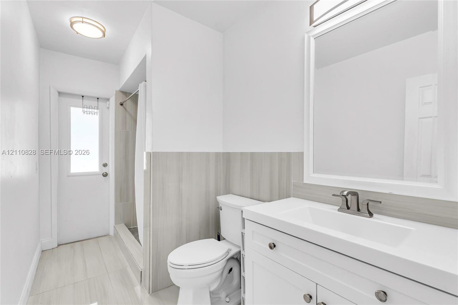 White vanity featuring an integrated sink, brushed metal faucet, and neutral-toned stone-finish backsplash, complemented by matching tile flooring, a tub-shower enclosure with tile surround, and a framed mirror