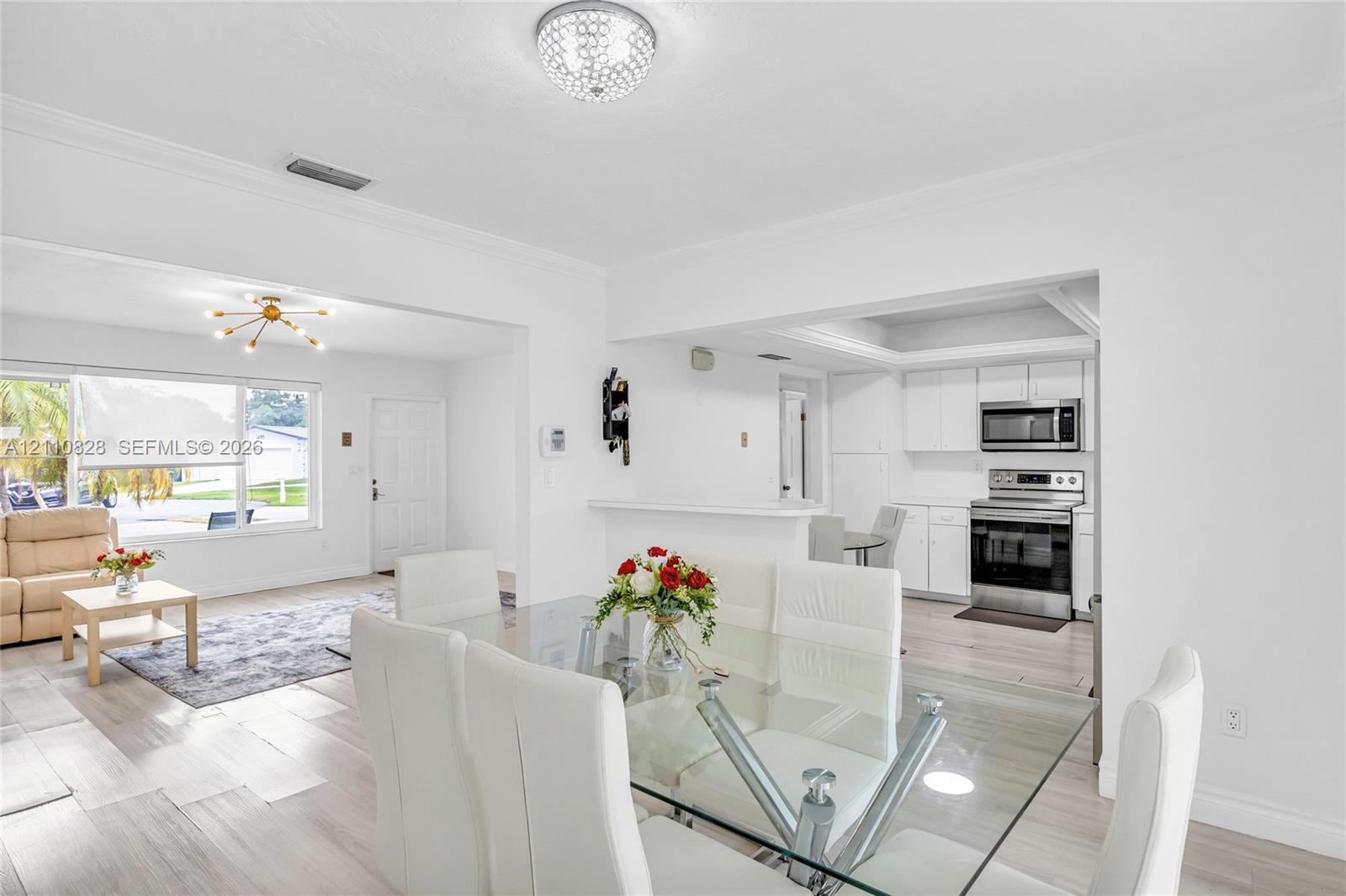 Open-concept interior featuring light wood-finish flooring, white shaker-style kitchen cabinetry, built-in stainless steel appliances, multi-arm chandelier lighting, and a tray ceiling