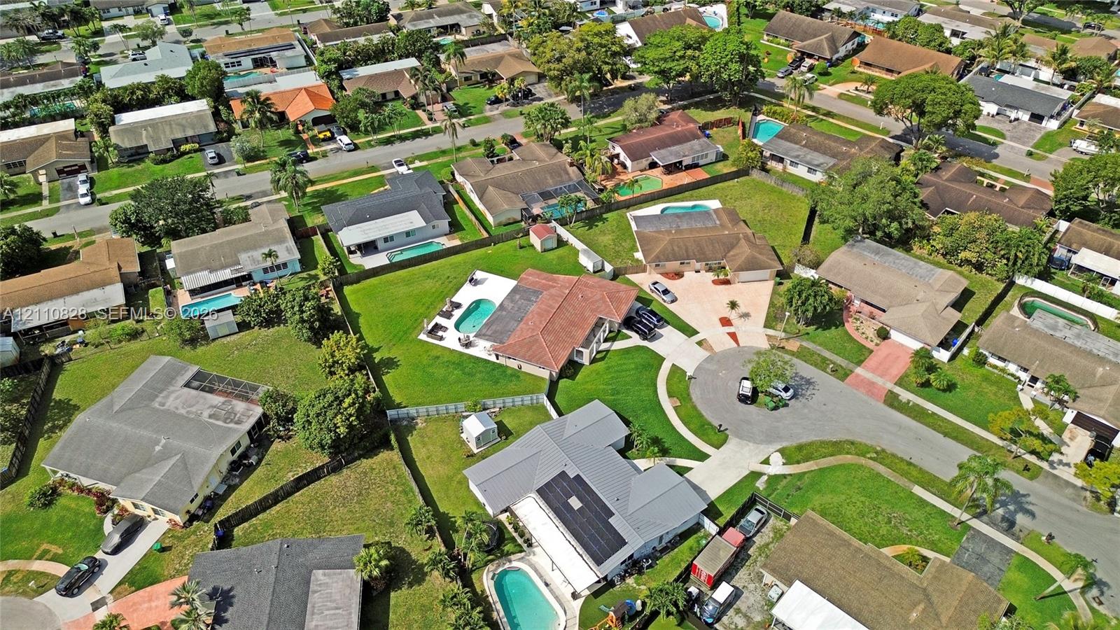 Aerial perspective of a residence featuring a clay tile roof, a rectangular swimming pool, a paved driveway, and a fenced-in grassy lawn