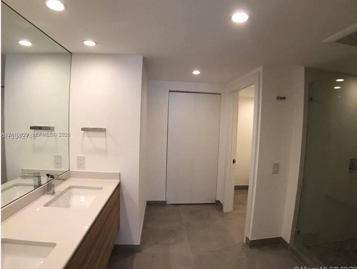 Floating wood-finish double vanity with stone countertop, recessed lighting, expansive wall-to-wall mirror, and glass-enclosed shower with tiled flooring
