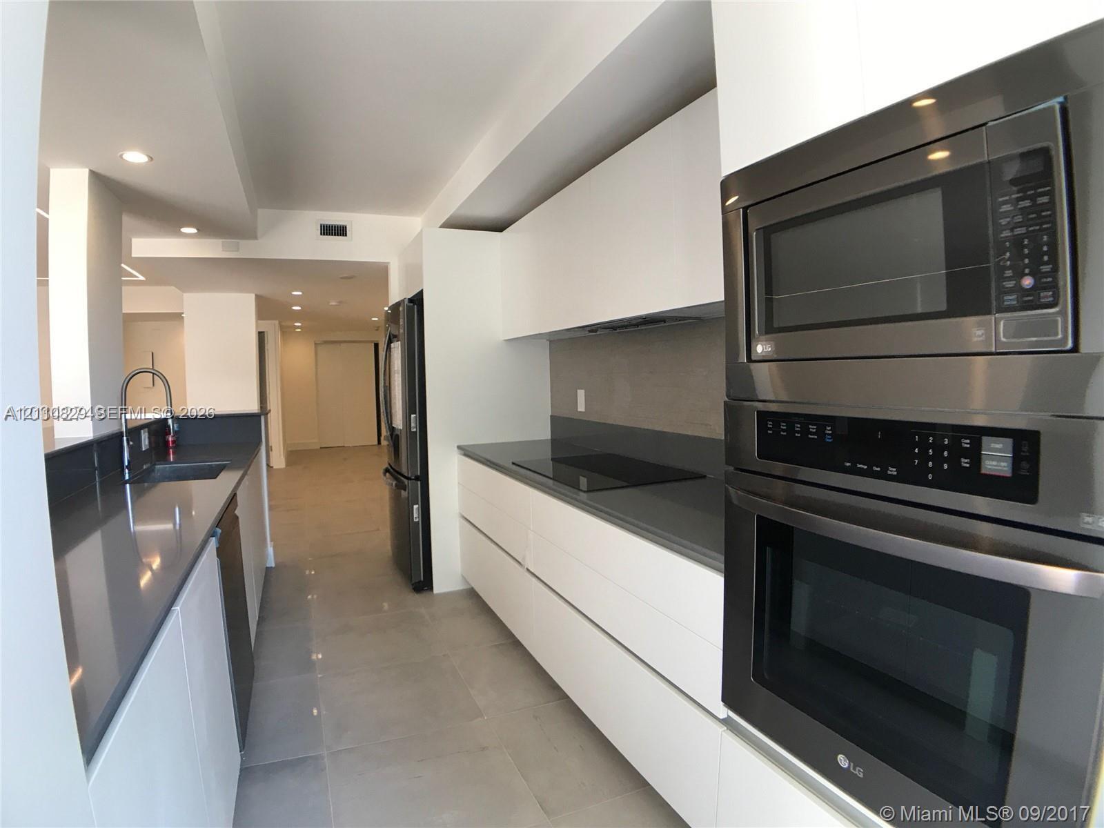 Kitchen featuring white flat-panel cabinetry, charcoal solid-surface countertops, stainless steel built-in double oven and microwave, recessed lighting, and large-format tile flooring