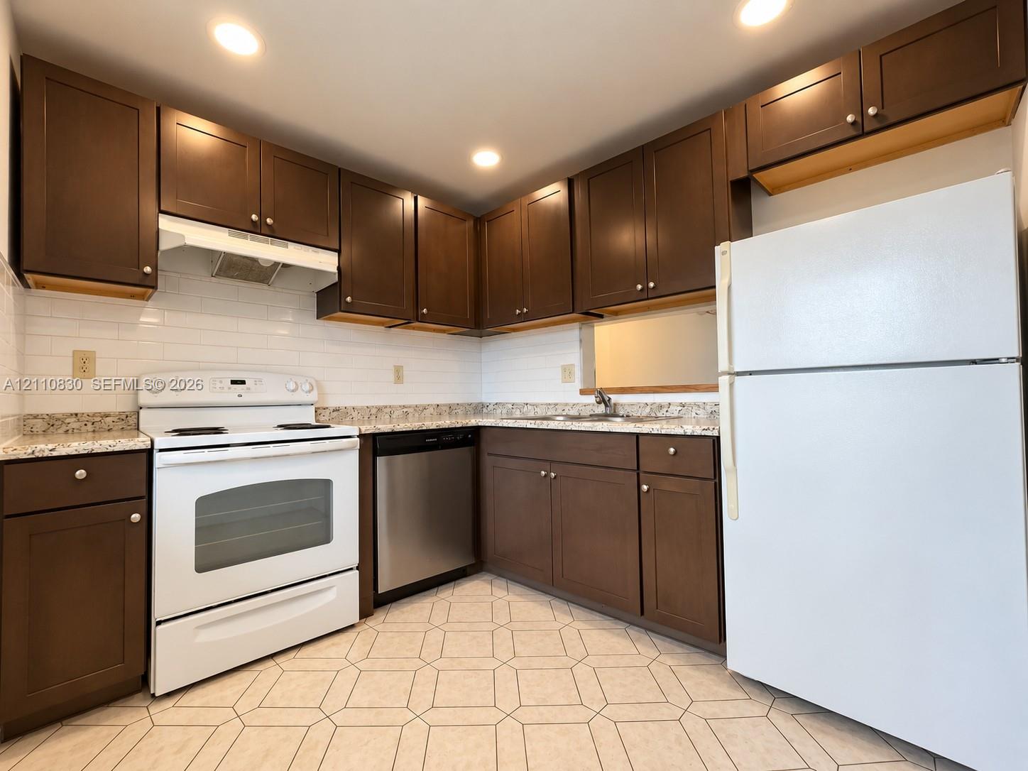 Spacious kitchen featuring rich wood cabinetry with ample storage, granite-style countertops, stainless steel appliances including a range and dishwasher, white refrigerator, and ceramic tile flooring throughout